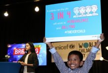 A boy holds up a placard that says If children ran the UN, 3 in 4 will work to end violence and bullying for every child. Behind him a girl presenter stands infront of a TV screen with the message Breaking News.