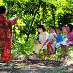 pmr_ashot_thumb_7 A teacher teaches four children sitting on a bench in the outdoors.